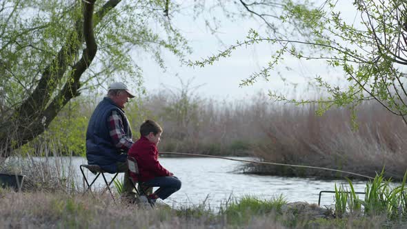 Anglers, an Elderly Man with Little Boy Catch Fish Using a Wooden Fishing Rod Sitting on Shore alt