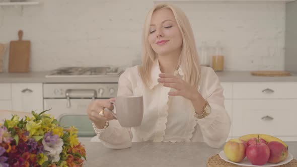 Close-up of a Blond Thoughtful Caucasian Lady Sitting at the Table with a Cup of Tea or Coffee alt