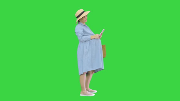 Happy Pregnant Woman in Blue Dress and Hat Using Her Smartphone on a Green Screen, Chroma Key. alt