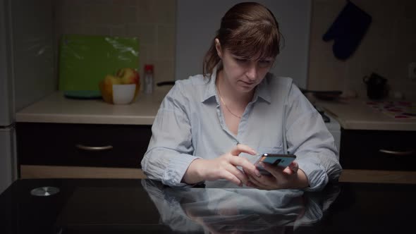 Serious Woman Sitting in the Kitchen at Night and Using the Phone