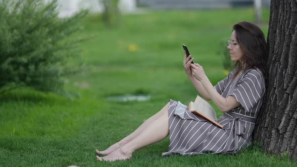 Lonely Adult Woman is Resting in Park at Summer with Book and Smartphone alt