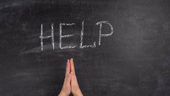 A woman's hand writes the phrase HELP in green chalk on a chalkboard