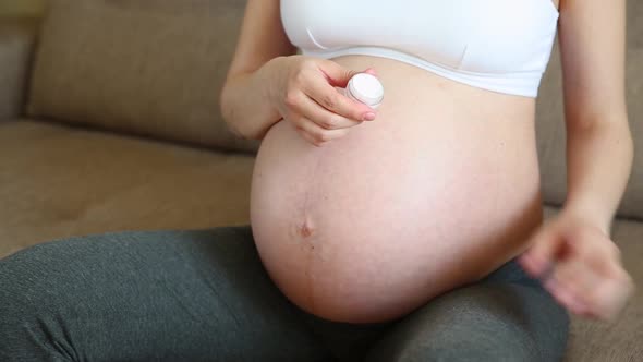 Woman Lubricates Moisturizes Pregnant Belly with Cream for Stretch Marks on Home Sofa alt