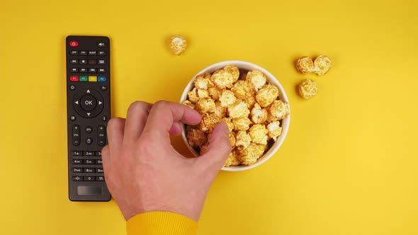 Male hands pick up crispy caramel popcorn from a white plate on a yellow background with a smart tv