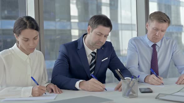 Middle Aged Businessman Writing Documents with His Assistants on Office Table alt