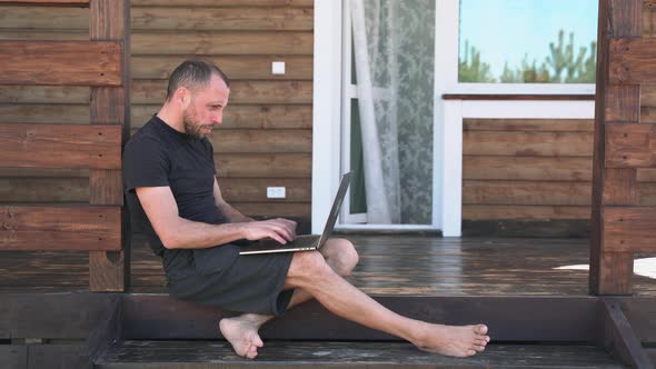 A Man in Shorts is Working on a Laptop Sitting on the Terrace Near the House A Businessman in a