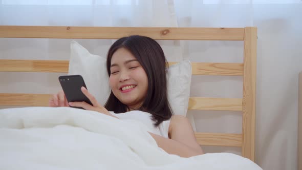 Young Asian woman using smartphone checking social media feeling happy smiling while lying on bed. alt
