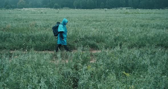 Woman in a Blue Raincoat and a Backpack Walks Through a Field with Grass
