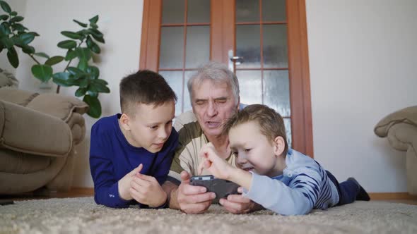 Two grandchildren are teaching grandfather to play their favorite toy on the tablet alt
