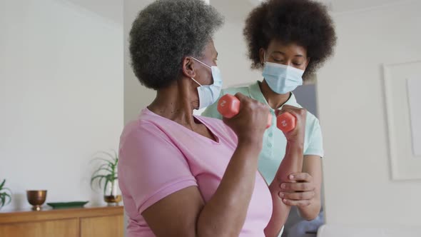 African american female physiotherapist wearing face mask helping senior female patient exercise alt