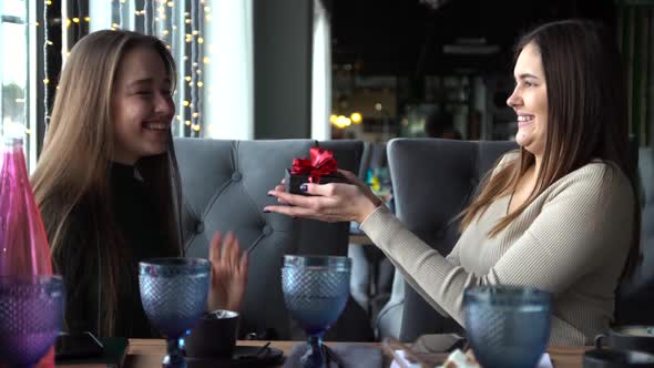 Beautiful Woman Giving Present To Her Surprised Friend While Sitting in Cafe.