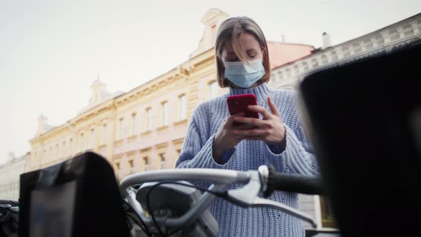 Tourist in medical mask take scooter or bike bicycle in sharing parking lot. alt