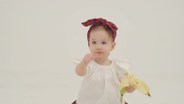 Little girl in plaid headband and skirt is eating big banana alt
