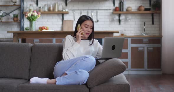 Young Asian Female Sitting in Cozy Sofa Talking on Phone at Home and Looking in Monitor of Her alt
