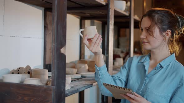 Portrait of Female Potter Checking Ceramics and Writing Information in Notebook in Workshop alt