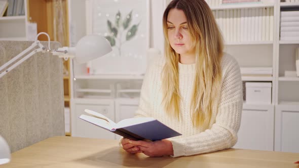 a Woman Sits at a Table and Reads a Book By the Light of a Table Lamp alt
