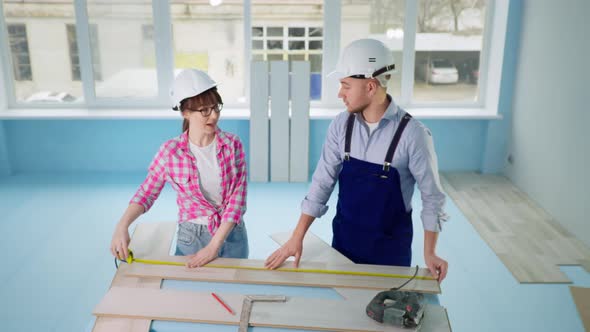 Woman and Man in Safety Helmets Measure Laminate Flooring with Tape Measure in Room During Redesign alt