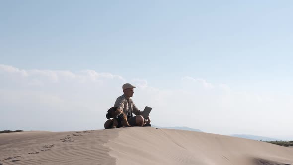 Male traveler sitting on sandy dune with dog and using laptop during vacation in sunny day alt