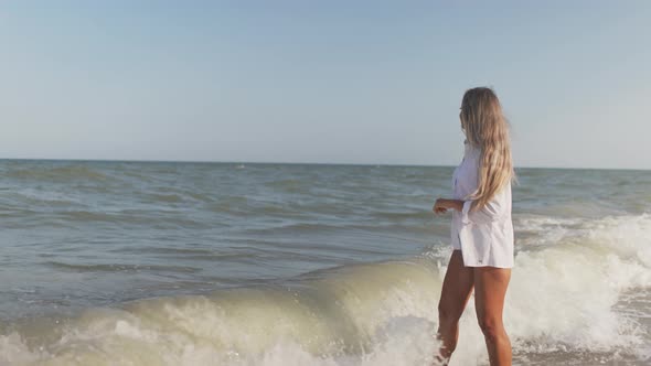 A Slender Girl in a Gentle Blue Swimsuit and Shirt Walks Along the Sandy Beach Near the Blue Sea alt