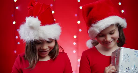 Two Cute Girls in Santa Hats Open Presents on Red Christmas Background alt