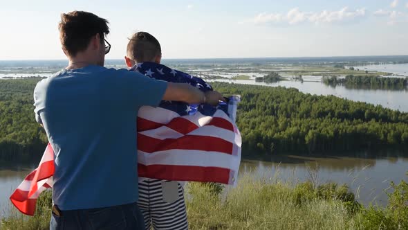 Father with American Flag Embraces His Son on River Bank