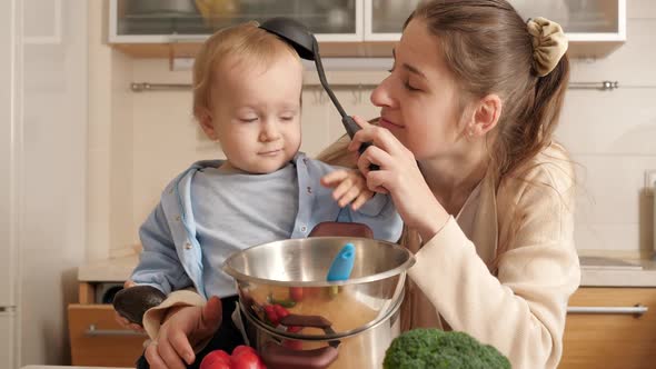 Happy Smiling Mother Playing with Ladle and Her Baby Son While Cooking on Kitchen alt