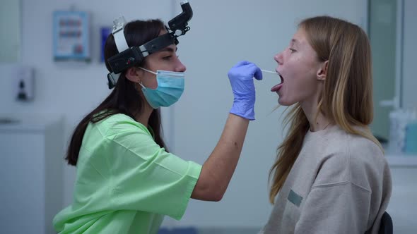 Side View Focused Woman with Disposable Spatula Checking Oral Cavity of Teenage Girl in Hospital alt