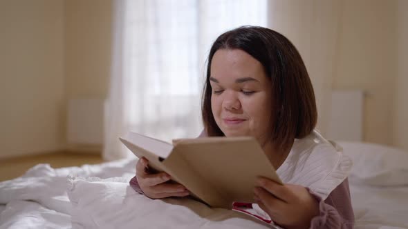 Young Happy Little Woman Reading Book Lying in Cozy Bed Smiling Looking Away alt