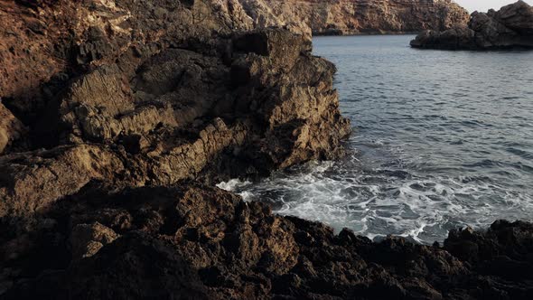 Rough sea waves crashing on a rocky shore. Dark brown cliffs by the sea in the sunset. alt