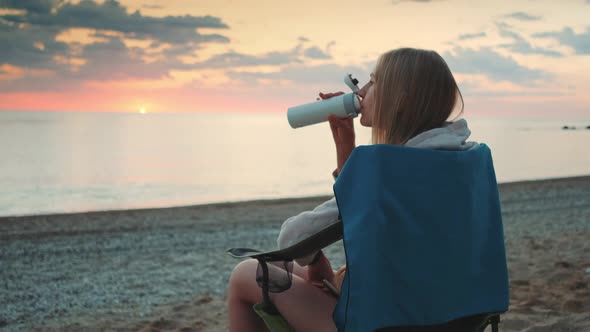 Young Woman Drinking From Thermos and Sitting on Camping Chairs on the Beach alt