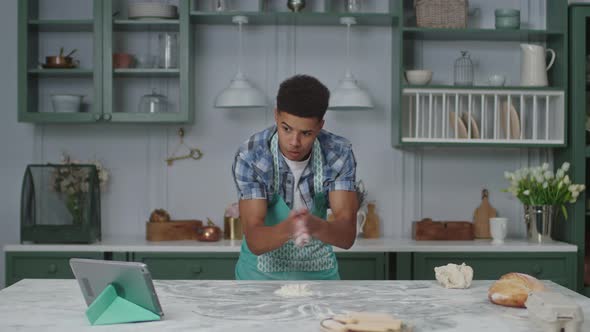 20s African American Man in Apron Kneading Dough with Flour Watching Tablet Computer alt