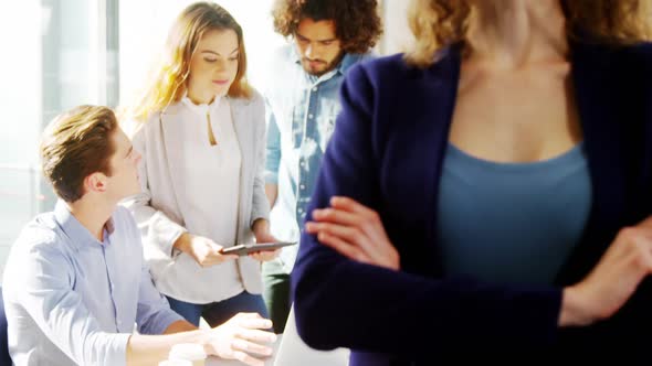 Woman standing with arms crossed while colleague discussing in background alt