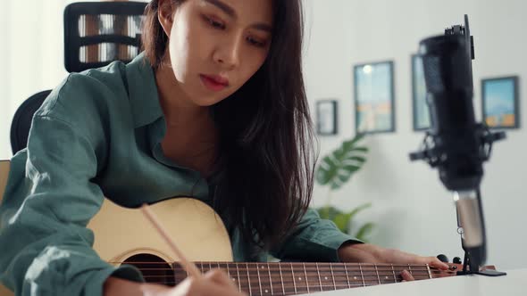 Happy asia woman songwriter play acoustic guitar listen song from smartphone think and write notes.
