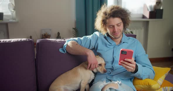 Man Sitting on Sofa in Livingroom Guy Using Smartphone Chatting Petting His Obedient Dog alt