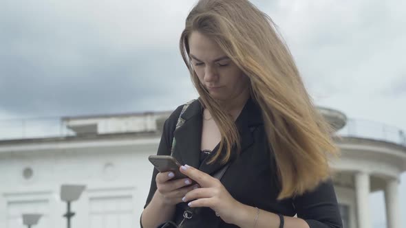 Concentrated Caucasian Woman Typing Message on Smartphone Outdoors. Portrait of Young Beautiful alt