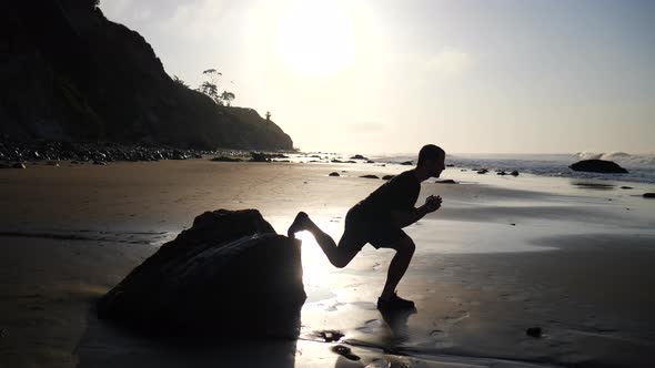 A strong muscular man doing leg lunges for a morning fitness workout at sunrise on a beach in Santa alt