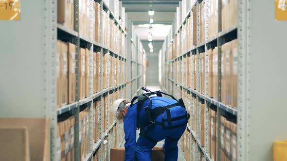 Warehouse Worker in Ergoskeleton is Storaging a Box Into the Shelf alt