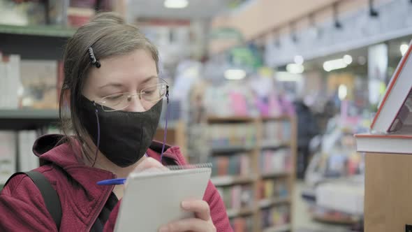 Woman in Glasses and Mask Writes Down Names of Books in the Store in a Notebook