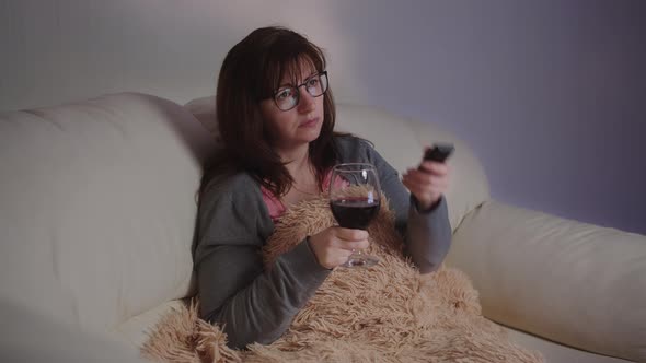 Sad Brunette Woman Relax at Home with a Glass of Wine on the Sofa and Watches Tv in the Evening alt