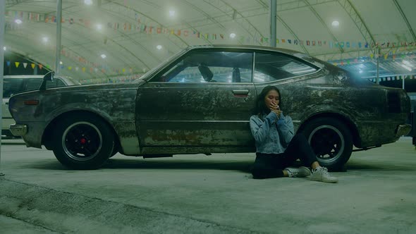 a Sad Thai Girl Sits Smoking Leaning Against a Rusty Car at Night alt