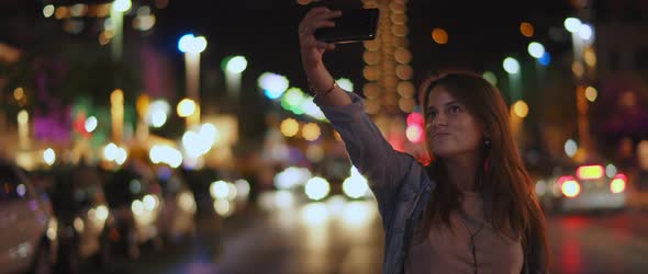 A young woman taking a selfie in the middle of the street, bokeh lights alt
