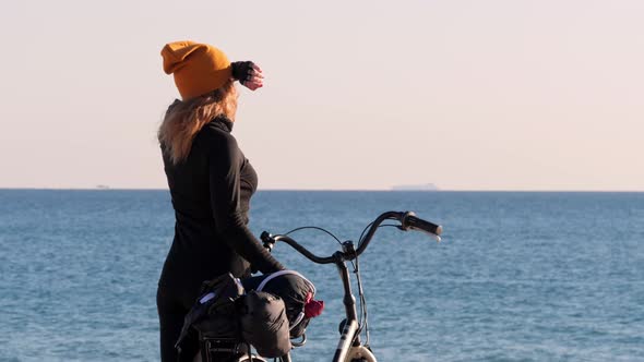 A Young Blonde Girl in Yellow Hat Holding the Her Bicycle and Looking at the Sea and Sunset