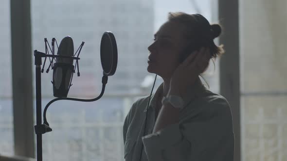 A Young Girl Emotionally Sings Into a Microphone in the Background of the Window alt