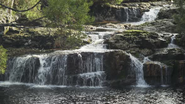 A small multi-level waterfall behind some branches on a sunny day, tight shot alt