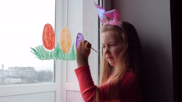 Cute little girl in bunny ears painting Easter eggs with green spring grass on window at home alt