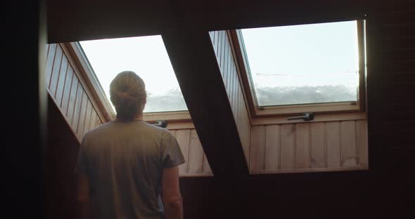 Back View of Man Standing Indoors in Wooden Interior Looking Out of Window alt