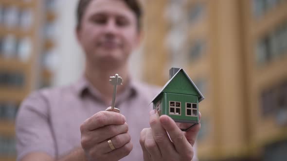 Closeup Toy House and Key in Male Hands with Blurred Happy Caucasian Man Smiling at Background alt