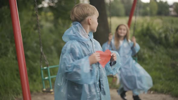 Portrait of Happy Cute Boy Standing with Red Paper Heart As Blurred Girl Swinging on Swings at the alt