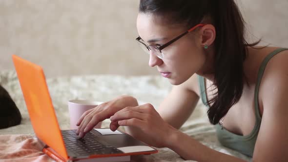 Young Attractive Girl Is Working on Her Laptop, Lying on the Bed at Home. A Young Cute Girl Is Lying