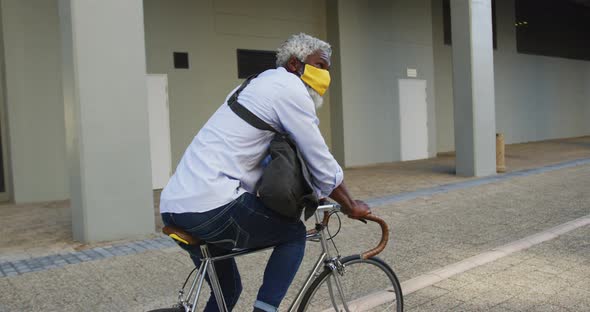 African american senior man wearing face mask riding bicycle in corporate park alt